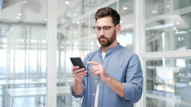 İşadamı, modern bir iş ofisindeki iş yerinde cep telefonunu kullanıyor. Gülümseyen yakışıklı işçi internette sohbet ediyor veya akıllı telefondan mesaj okuyor ve kameraya bakıyor