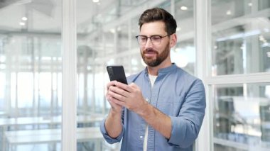 Tatmin olmuş mutlu iş adamı, modern iş ofisindeki işyerinde dururken cep telefonundan harika haberler aldı. Gülümseyen yakışıklı girişimci akıllı telefondan olumlu bir mesaj okur.