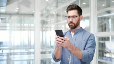 Endişeli işadamı, modern bir iş ofisinde işyerinde dururken cep telefonundan kötü haberler okuyor. Sinirli erkek işçi, akıllı telefondaki olumsuz bilgileri incelerken üzgün.