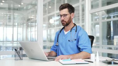 Mavi ceketli kendine güvenen doktor hastane ofisinde dizüstü bilgisayarla konuşuyor. Erkek doktor konferansta uzaktan iletişim kurar ya da bir hastayla çevrimiçi istişarede bulunur.