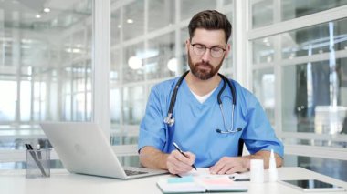 Mavi ceketli gülümseyen doktorun portresi modern hastane kliniğinin çalışma masasında kameraya bakarken mutlu sakallı, profesyonel doktor büroda evrak işleriyle uğraşıyor.