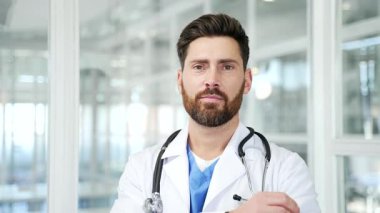 Modern hastane kliniğinde kameraya bakarken beyaz önlüklü, gülümseyen yakışıklı doktorun portresi. Ofis ortamında poz veren mutlu, profesyonel bir tıp doktorunun vesikalığı. Kapat.