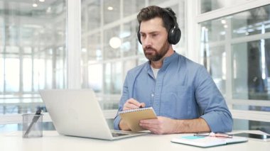 Kulaklıklı bir iş adamı bilgisayarını kullanarak video konferans izliyor iş yerindeki iş yerinde oturan not defterine not alıyor. Uzaktan çevrimiçi eğitim seminerini dinleyen odaklanmış işçi