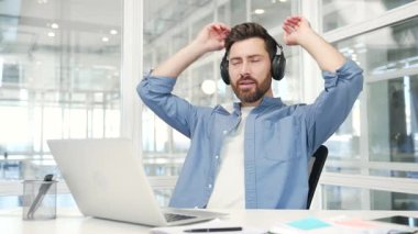 İş yerinde bir iş yerinde otururken, kulaklıklı mutlu bir işadamı iş arasında müzik dinliyor. Neşeli işçi rahatlar, gözleri kapalı dinlenir ellerini başının arkasına koyar.