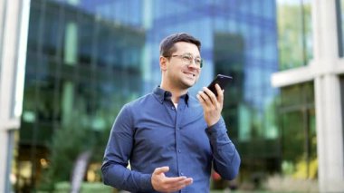 İş adamı, akıllı telefondan sesli mesaj kaydediyor. Telefonu ağzına yakın tutuyor. Ofis binasının yanındaki sokakta yürüyor. Adam konuşmalarını mobil uygulama üzerinden dikte ediyor, hoparlörle sohbet ediyor.