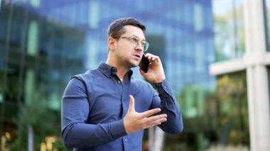 Öfkeli hoşnutsuz işadamı, modern ofis binasının yanındaki caddede telefonla konuşuyor. Heyecanlı adam duygularını ifade ediyor, stresli iş durumlarıyla uğraşıyor, çatışma çözülüyor.
