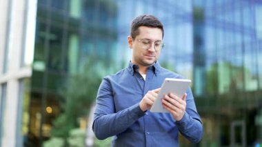 Kendine güvenen işadamı, modern bir ofis binasının yanında sokakta dikilirken dijital tablet kullanıyor. İnternette ciddi yakışıklı erkek işçiler sohbet ediyor, internette geziniyor ya da mesajlar yazıyor. Kapat.