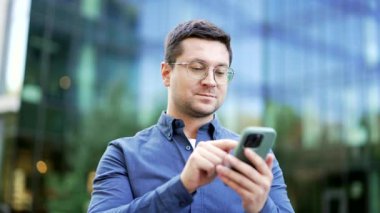 Gülümseyen işadamı modern bir ofis binasının yanında caddede dururken cep telefonu kullanıyor. Çevrimiçi yakışıklı erkek sohbetleri internette geziniyor ya da akıllı telefondan mesajlar yazıyor. Kapat.