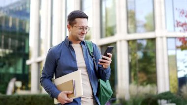 Gülümseyen erkek öğrenci üniversite binası yakınlarında kampüste yürürken cep telefonu kullanıyor. Sırt çantası, kitapları ve ders kitapları olan yakışıklı bir adam arkadaşlarıyla internette sohbet ediyor, internette geziniyor, mesajlar yazıyor.