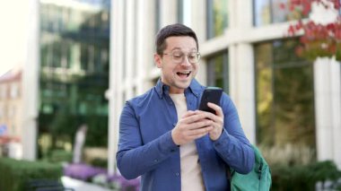 Mutlu, heyecanlı erkek öğrenci, üniversite binasının yakınındaki kampüs alanında beklerken cep telefonundan harika haberler aldı. Gülümseyen şaşkın yakışıklı adam olumlu bir mesaj okuyor.