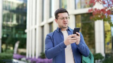 Endişeli erkek öğrenci üniversite binasının yanında kampüs dışında dururken cep telefonundan kötü haberler okuyor. Sinirli ve stresli yakışıklı adam akıllı telefondan gelen olumsuz mesaja baktığı için üzgün.