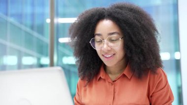 Gülümseyen Afro-Amerikan iş kadını iş yerindeki masasında dizüstü bilgisayarda oturuyor. Siyahi kadın çalışan bir projede bilgisayar üzerinde çalışıyor ya da bir işle meşgul. Kapat.