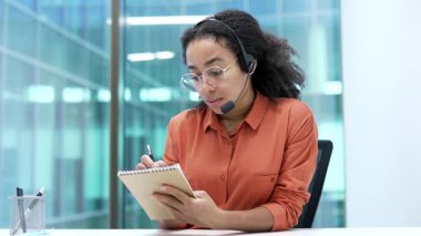 Webcam görüntüsü. Afrika kökenli Amerikalı iş kadını kulaklıkla video konferans izliyor ofisteki kameraya bakarak notlar alıyor. İş eğitimi alan siyahi kadın uzaktan iletişim kuruyor.