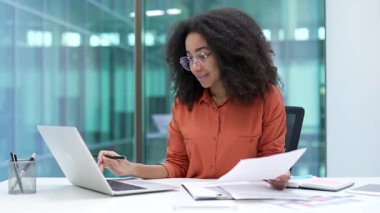 Modern iş ofisindeki çalışma masasında oturan gülümseyen Afrikalı Amerikalı finansçının portresi. Gözlüklü mutlu iş kadını bilgisayarda çalışıyor, evrak işleriyle meşgul ve kameraya bakıyor.