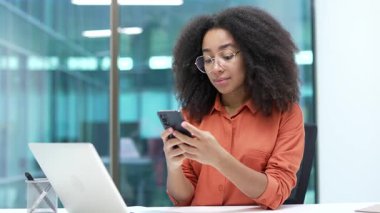 Genç Afro-Amerikan iş kadını iş yerindeki masasında telefon kullanıyor. Siyahi kadın çalışan mobil uygulama üzerinde çalışıyor, daktilo mesajı okuyor, internette sohbet ediyor veya internette geziniyor.