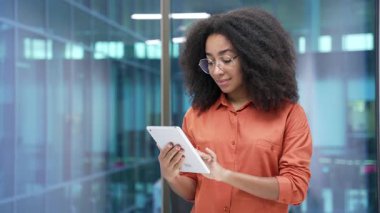 Modern cam iş ofisinde işyerinde duran gözlüklü, gülümseyen Afro-Amerikan iş kadını portresi. Elinde dijital tabletle kameraya poz veren mutlu zenci kadın çalışan.