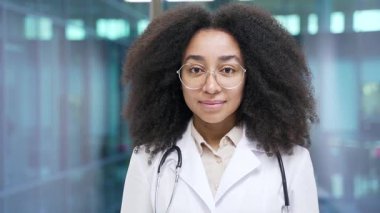 Hastane kliniğinde kameraya bakan beyaz önlüklü ve gözlüklü gülümseyen Afro-Amerikan kadın doktor portresi. Kafasından vurulmuş mutlu siyahi kadın doktor ofiste duruyor. Kapatın.