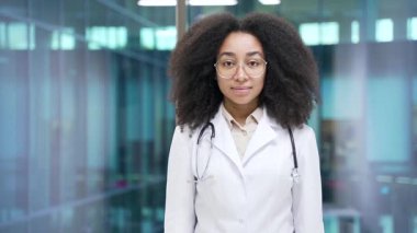 Hastane kliniğindeki kameraya bakan beyaz önlüklü, ciddi bir Afrikalı Amerikalı kadın doktorun portresi. Kendine güvenen siyahi kadın tıp doktoru modern ofiste yan yana duruyor.