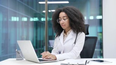 Hastane kliniğindeki çalışma masasında oturan beyaz önlüklü ciddi bir Afrikalı Amerikalı kadın doktor portresi. Modern ofiste kameraya bakan gözlüklü siyahi doktor kadın.