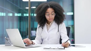 Afro-amerikan kadın doktor, hastane kliniğindeki iş yerinde dizüstü bilgisayarında daktilo kullanarak reçete yazıyor. Beyaz önlüklü genç siyahi kadın doktor ofiste çalışıyor.