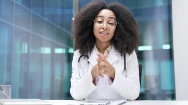 Webcam görüntüsü. Afro-Amerikan kadın doktor, hastane kliniğindeki kameraya bakarak görüntülü konuşuyor. Tıp doktoru, ofisinde oturan bir hastayla uzaktan görüşüyor.