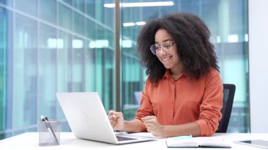 Genç, mutlu, Afro-Amerikan iş kadını iş yerinde oturmuş laptopta iyi haberler okuyor. Gülümseyen siyah kadın seviniyor olumlu mesaj bildirimi