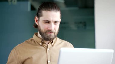 İş yerindeki bilgisayarında çalışan gülümseyen bir iş adamı. Yakışıklı sakallı erkek işçi bilgisayarda yazı yazıyor, müşteriye mesaj atıyor, internette sohbet ediyor ya da bir projeyle meşgul. Kapat.