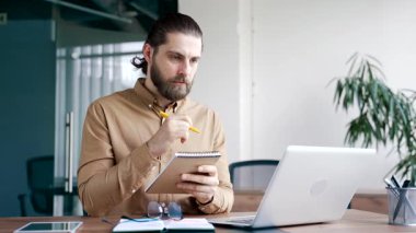 İş adamı bilgisayar kullanarak video konferansı izliyor. İş yerindeki not defterine not alıyor. Uzaktan eğitim, kurs, seminer dinleyen odaklanmış bir işçi. Yeteneğini kullan.