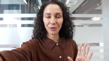 İş yerindeki bir iş yerinde cep telefonuyla konuşan genç, gülümseyen bir iş kadını. Mutlu kadın çalışan kameraya bakıyor selfie çekiyor, selamlıyor, el sallıyor, uzaktan iletişim kuruyor.