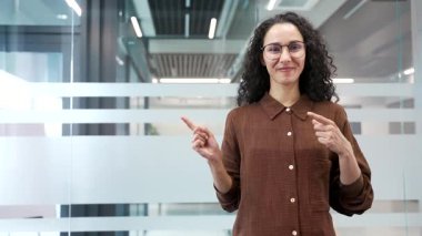 Gülümseyen iş kadını cam ofiste dururken mesaj yazmak için boş yeri işaret ediyor. Mutlu bayan çalışan kameraya bakar. Reklam şablonu. Terfi için boşluk kopyala