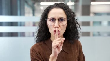 Genç iş kadını, modern iş ofisindeki işyerinde dururken işaret parmağıyla dudaklarını işaret ediyor. Kadın işçi sessizlik jesti yapıyor. Sessiz olun, ses çıkarmayın, sessiz olun diyor. Kapat.
