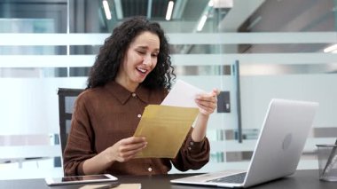 Mektubu büyük haberlerle okuyan mutlu iş kadını, modern bir iş yerinde otururken başarıyı kutluyor. Gülümseyen heyecanlı bayan çalışanımız hoş bir tebligattan memnun.