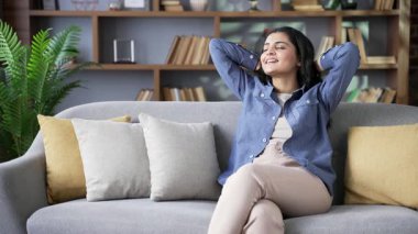 Evdeki oturma odasında rahat bir kanepede otururken gözleri kapalı ve elleri başının arkasında rahatlayan genç bir kadın. Sakin, güzel bir kadın, dinleniyor, rahatlamış hissediyor.