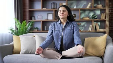 Nilüfer pozisyonunda meditasyon yapan genç bir kadın evdeki oturma odasında kanepede oturuyor. Sakin, mutlu kadın yoga yapıyor, gözleri kapalı, derin nefes alıyor, rahat bir koltukta rahat bir atmosferde dinleniyor.