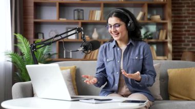 Kendine güvenen genç bir kadın ofisteki bilgisayarı ve mikrofonu kullanarak podcast yapıyor. Kadın vlogger etkileyicisi çevrimiçi yayın yapıyor, röportaj veya webinar, video çağrısı ile uzaktan konuşuyor