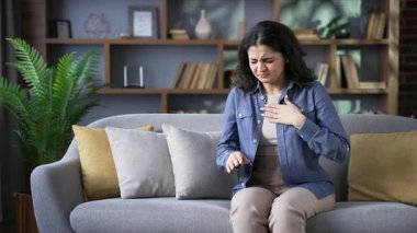 Evdeki oturma odasında kanepede oturan hasta genç kadın öksürüyor. Sağlıksız yetişkin dişi kendini kötü hissediyor, elini göğsüne dayıyor. Kız nezle, virüs, grip, bronşit veya zatürree olmuş.