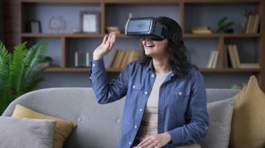 Video görüşmesi için VR gözlüğü kullanan ve evdeki oturma odasındaki kanepede oturmuş sanal gerçeklik simülatöründe sohbet eden genç bir kadın. Mutlu yetişkin kadın uzaktan iletişim kuruyor, online bir toplantısı var.