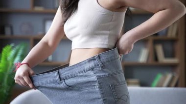 Genç, yetişkin bir kadın, evinde oturan büyük beden kot pantolon giyerek kilo verme başarısını sergiliyor. Fitness kavramı, kişisel başarı, sağlık dönüşümü, yaşam tarzı değişimi.