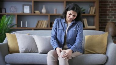 Mutsuz genç kadın evdeki oturma odasında kanepede otururken şiddetli diz eklem ağrısı çekiyor. Üzgün yetişkin kadınlar bacak kaslarına masaj yapar, yürüyemez, hastalık, kramp veya romatizma geçiremez.