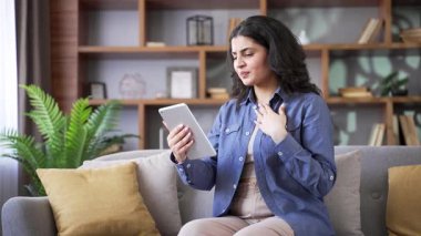 Hasta kadın evdeki oturma odasındaki kanepede dijital tablet kullanan bir doktorla görüntülü konuşma yapıyor. Kalp rahatsızlığı olan yetişkin bir kadın sorunlarını anlatıyor, hastalığından şikayet ediyor.