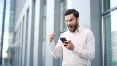 Mutlu iş adamı modern ofis binasının yanındaki caddede duran cep telefonundan harika haberler aldı. Şok olmuş gülümseyen gözlüklü işçi akıllı telefondan olumlu mesaj okuyor.