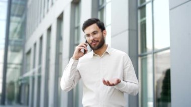 Ofis binasının yanındaki sokakta cep telefonuyla konuşan gülümseyen bir işadamı. Yakışıklı girişimci iş ortağı ya da müşterisiyle iletişim kurar. Profesyonel adam akıllı telefondan konuşuyor.
