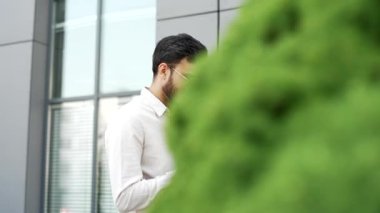 Modern ofis binasının yanındaki yeşil ağaçların yanında yürüyen cep telefonuna bakan işadamlarının yan görüntüsü. Girişimci akıllı telefona göz atıyor, internette sohbet ediyor ya da mesajları okuyor. Kapat.