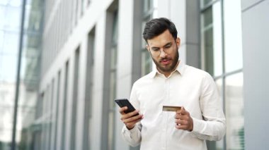 İşadamı, iş yeri binasının yanında caddede dururken kredi kartı ve cep telefonu kullanarak online alışveriş yapıyor. Girişimci faturaları öder, internete para harcar, e-ticaret e-alışveriş yapar.