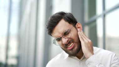 İş adamı, bir ofis binasının yakınında dikilirken kulak ağrısından muzdarip. Adam parmaklarıyla kulağına dokunuyor. Amcası, kiremitleri, egzaması, kulak hastalıkları ya da kulak iltihabı var. Kulak ağrısı. Kapat. 