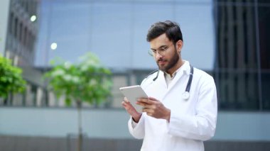 Hastane binasının önünde dikilirken dijital tablet kullanan beyaz önlüklü kendine güvenen bir doktor. Ciddi yakışıklı doktor gözlüklü bir doktor hastayla internette sohbet ediyor ya da uygulama üzerinde çalışıyor.