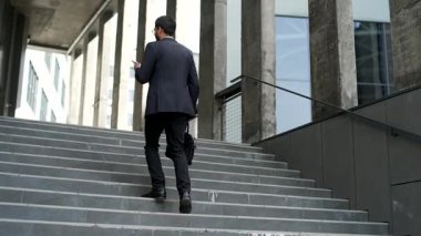 İş adamı elinde akıllı telefonuyla ofis binasının merdivenlerinden çıkıyor. Resmi takım elbiseli, evrak çantalı, kendine güvenen girişimci şehir merkezindeki telefonları kontrol ediyor. Profesyonel bir iş. Arka görünüm