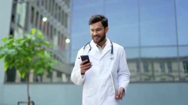 Beyaz önlüklü gülümseyen doktor hastane binasının yanında cep telefonu kullanıyor. Yakışıklı doktor akıllı telefonu kontrol eder, mesajları okur, internette gezinir ya da hastalarla sohbet eder.