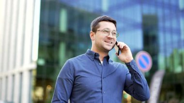 Ofis binasının yakınında yürürken cep telefonuyla konuşan gülümseyen bir işadamı. Yakışıklı erkek iş ortağı ya da müşterisiyle iletişim kurar. Profesyonel adam akıllı telefon kullanarak konuşuyor.