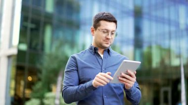Ofis binasının yanındaki sokakta elinde tablet bulunan gülümseyen iş adamının portresi. Mavi gömlekli mutlu yakışıklı erkek kameraya bakıyor. Kendine güvenen bir iş adamı dışarıda poz veriyor.